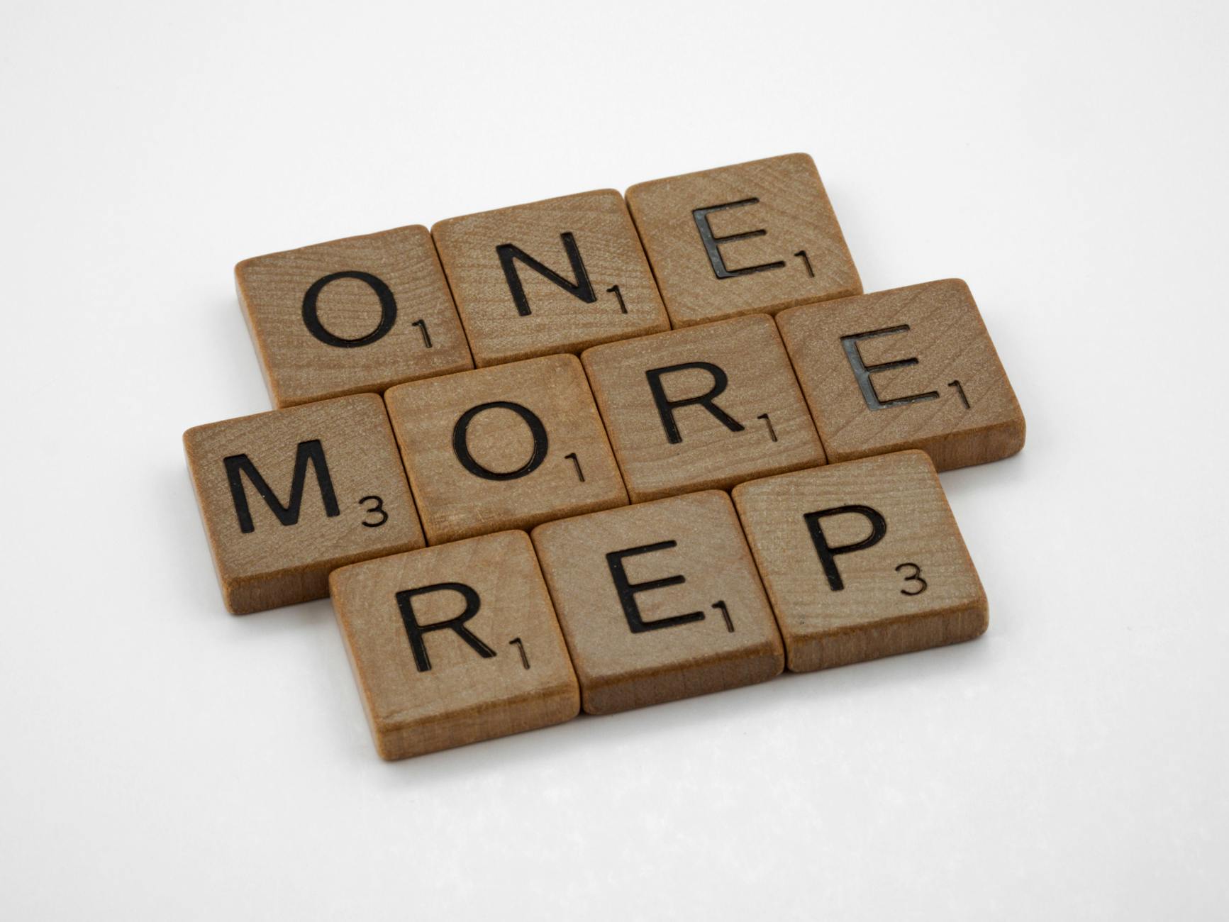 Wooden Scrabble tiles arranged to spell out the phrase "ONE MORE REP" on a plain white background.