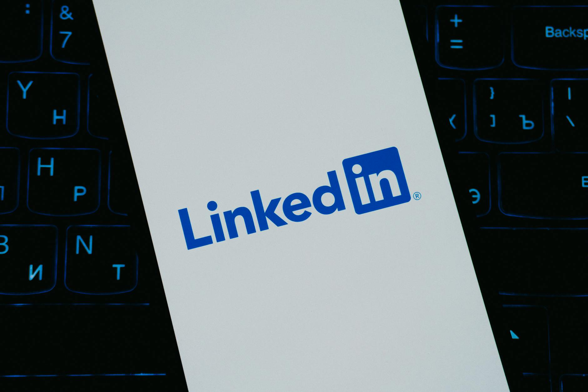 A smartphone screen displays the LinkedIn logo in blue and white, placed over a dark computer keyboard in the background. A smartphone screen displays the LinkedIn logo in blue and white, placed over a dark computer keyboard in the background.