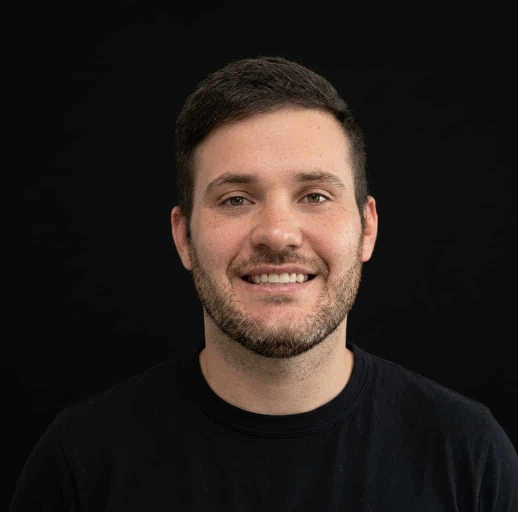 A man with short dark hair and a trimmed beard smiles at the camera. He is wearing a plain black T-shirt and is posed in front of a solid black background.