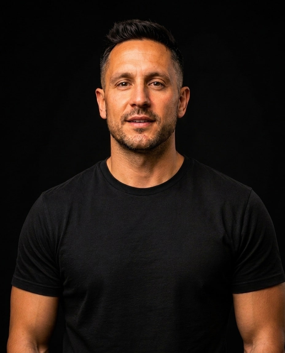 A man with short dark hair and facial stubble wears a plain black t-shirt and stands against a black background, looking directly at the camera with a neutral expression.